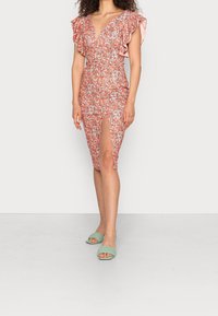 Robe florale avec des manches à volants, décolleté en V et fente sur le côté. Présente un motif vibrant orange et rose. Portée avec des sandales ouvertes à bout vert en tressé.