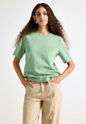 Kvinde med langt krøllet hår iført lysegrøn kortærmet skjorte og beige bukser, stående med hænderne i lommerne mod en hvid baggrund.