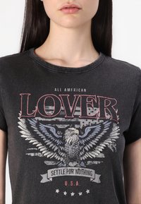 Žena v černém tričku s grafikou orla, na pozadí americká vlajka a text "All American Lover, Settle For Nothing, U.S.A."