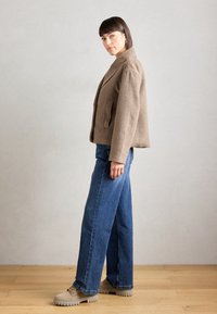 Veste en laine marron avec un col et des poches à rabat, associée à un jean bleu à jambes larges et des chaussures à semelles épaisses beiges.