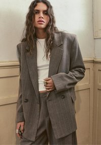 Donna con capelli lunghi e mossi che indossa un completo marrone a righe oversize e una camicia bianca corta, in piedi contro una parete beige a pannelli.