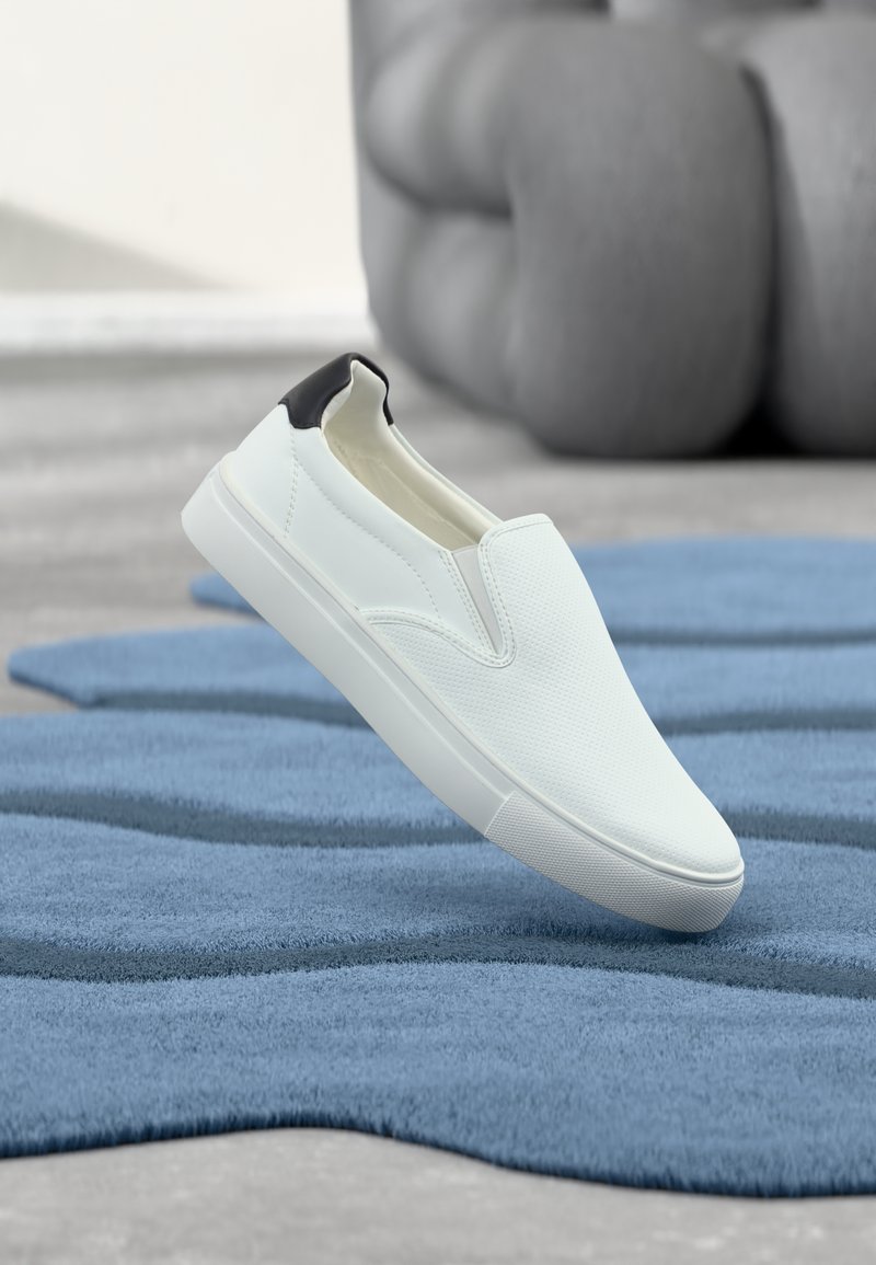 Chaussure blanche sans lacets avec une surface texturée, talon noir contrastant, semelle en caoutchouc plate, et un design épuré, posée sur un tapis bleu.