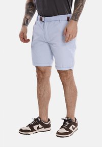 Pantaloni shorts azzurri chiari realizzati in tessuto morbido, dotati di una cintura intrecciata, tasche frontali e orli arrotolati. Indossati con sneaker marroni e bianche.