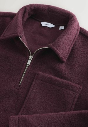 Vinröd fleecepullöver med dragkedja, en stor lappficka och en strukturerad krage. Mjuk textur och varmt material.