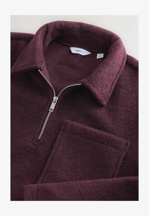 Vinröd fleecepullöver med dragkedja, en stor lappficka och en strukturerad krage. Mjuk textur och varmt material.