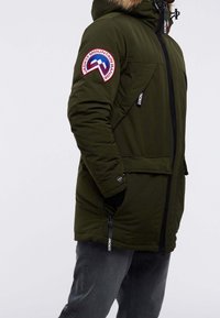 Olive green puffer jacket with a fur-lined hood, front zipper, and two side pockets; features logo patch on the left sleeve.