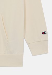 Beige kapucnis pulóver kenguruzsebbel, bordázott mandzsettával, és piros, valamint kék hímzett logóval a bal ujjon. Puha, sima textúra.