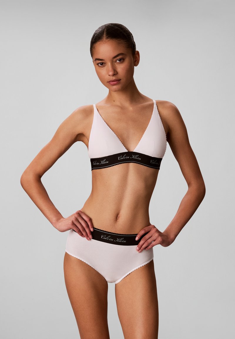 Γυναίκα που φοράει ανοιχτό ροζ σουτιέν Calvin Klein και ταιριαστό εσώρουχο, στέκεται με τα χέρια στη λεκάνη μπροστά σε απλό γκρι φόντο.