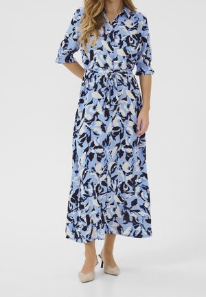 Frau trägt ein midi-Kleid mit abstraktem blau-schwarz-weißem Muster, Knopfleiste, gebundener Taille und beigen spitzen Absätzen.