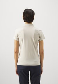 Šviesiai bežinė trumpomis rankovėmis polo marškinėliai su minkšta texture ir klasikinėmis apykaklėmis, dėvimi su tamsiai mėlynomis kelnėmis, matomi iš nugaros.