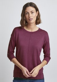 Burgundy stickad tröja med rund halsringning och trekvartsärmar. Har dekorativa knappar längs ärmen. Strukturerad tyg.