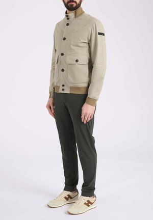 Uomo che indossa una giacca beige con bottoni e grandi tasche frontali, pantaloni scuri e scarpe da ginnastica beige con dettagli marroni, in piedi su sfondo semplice.