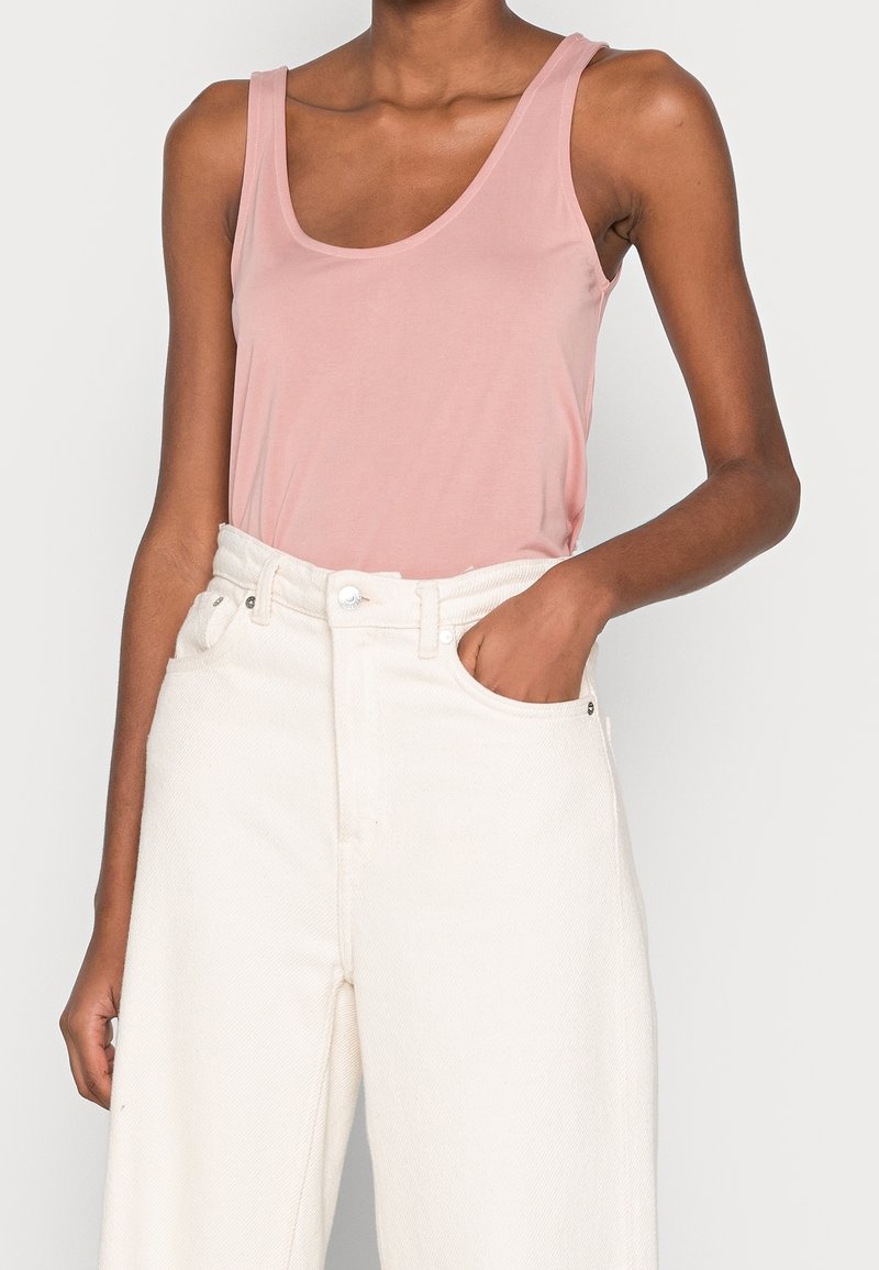 Femme portant un haut sans manches rose clair rentré dans un pantalon blanc ample à taille haute, avec la main droite dans la poche.
