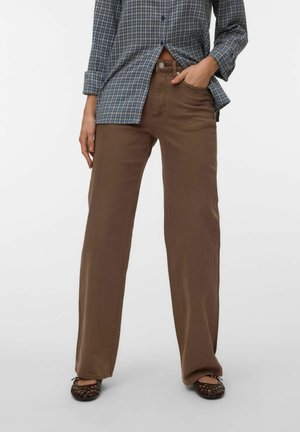 Naine kannab pruune laiade säärtega pükse, tume kootud korterjalanõusid ja sinist ruudulist nööbitavat särki, mille varrukad on üles keeratud. Üks käsi taskus.