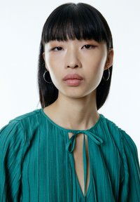 Teal geplooide blouse met een strikhals, voorzien van lange mouwen en een subtiele glans. Model draagt zilveren hoepelringen, neutrale make-up en steil zwart haar.