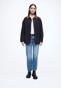 Casaco azul-marinho com gola em bombazine castanha, fecho de pressão frontal, bolsos laterais, usado sobre um suéter branco e jeans azul claro. Botas pretas.