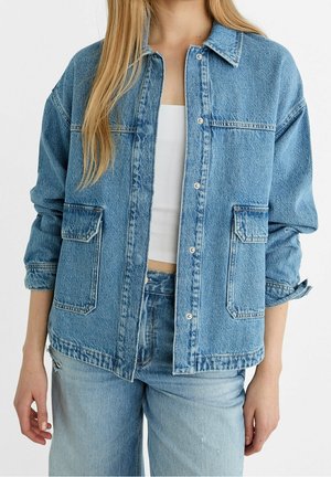 Femme portant une veste en denim bleu clair avec boutons-pression, grandes poches à rabat, manches retroussées, associée à un jean assorti et un haut blanc court.