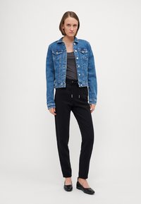 Veste en jean de couleur bleu moyen avec fermeture à boutons, deux poches poitrine et une coupe ajustée. Associée à un pantalon noir ajusté et des chaussures plates.