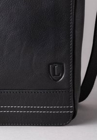Black leather bag featuring textured surface, embossed logo, contrasting stitching, and a sleek design. Accents include a shield shape.