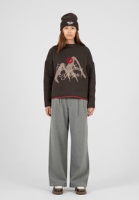 Brązowy sweter z grafiką gór i napisem, w połączeniu z szarymi spodniami o szerokich nogawkach. Dodatki to prążkowana czapka i brązowe buty.