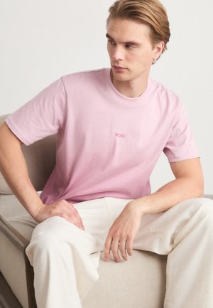 T-shirt en coton rose à manches courtes, avec un logo subtil dans une teinte plus claire. Coupe décontractée, associée à un pantalon de couleur crème.