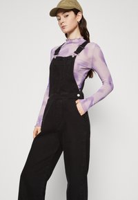 Black denim overalls with adjustable straps, a front pocket, and a loose fit, paired with a sheer purple long-sleeve top and a beige cap.