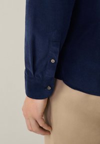 Camisa de franela de color azul marino con mangas y cierre de botón, que presenta una tela texturizada y un leve brillo, junto a una tela de color beige claro en la parte inferior.
