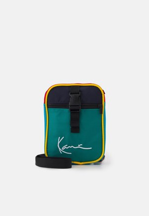Petit sac bandoulière rectangulaire en bleu sarcelle, bleu marine, rouge et jaune avec sangle noire, boucle à l'avant, poche zippée et logo blanc en écriture manuscrite.