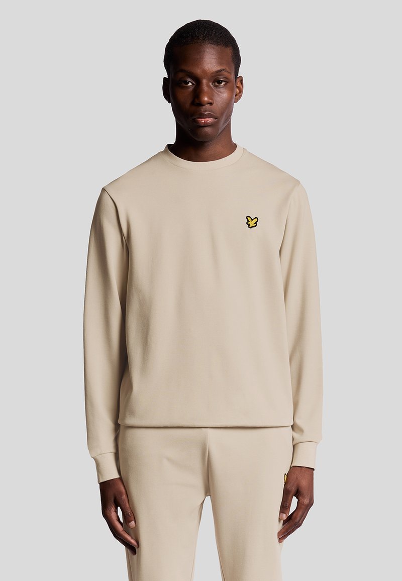 Beige sweatshirt met lange mouwen in een gladde stof, met een geel logo op de borst. Eenvoudige ronde hals en rechte zoom.