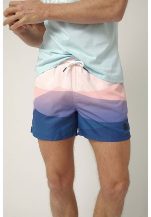 Hombre con pantalones de baño en bloques de colores pastel con rayas horizontales en azul claro, rosa, púrpura y azul oscuro, combinado con una camisa azul claro.