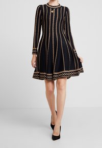 Robe noire en tissu texturé, avec manches longues, jupe évasée et motif chaîne dorée le long de lignes verticales.