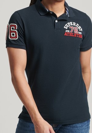 Mężczyzna w granatowej koszulce polo z białym i czerwonym logo "Superdry 76 Athletics" na piersi oraz numerem "76" na lewym rękawie.