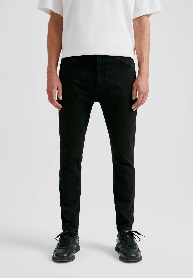Homme portant un jean noir slim, des baskets noires et un t-shirt blanc à manches courtes, debout devant un fond uni.