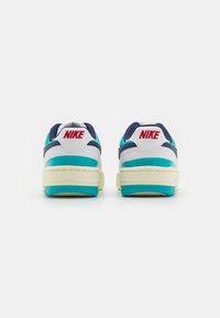 Nike-sneakers har en vit ovandel med marinblå och teal detaljer samt en röd logotyp, textilier med struktur och en kraftig krämfärgad sula med blå detaljer.