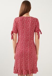 Robe fleurie rouge avec manches courtes et détails à nœud. Caractérisée par un ourlet volants et une silhouette ajustée avec un tissu à motifs.