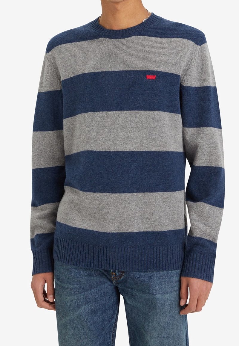 Pull rayé bleu marine et gris, en tissu doux. Présente un col et des poignets côtelés, avec un petit logo de marque rouge sur la poitrine.