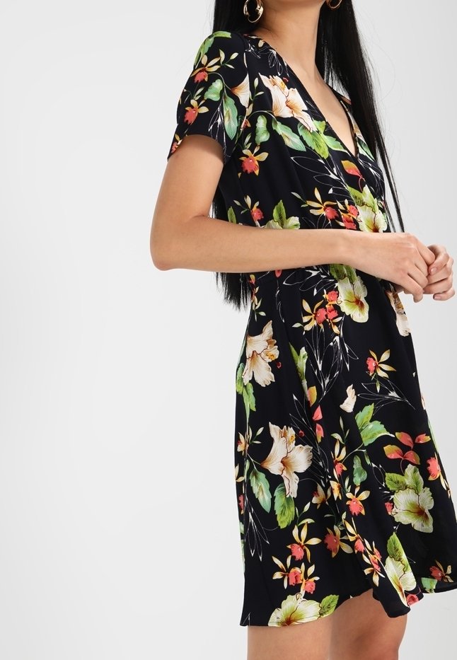 Kvinna klädd i en svart blommig wrapklänning med korta ärmar, prydd med stora gröna, vita och rosa blommor, som står framför en enkel bakgrund.