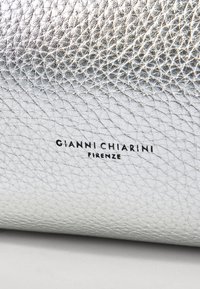 Zilverkleurig gestructureerd leer met een geometrisch patroon, met zwart merktekst: "GIANNI CHIARINI FIRENZE" duidelijk zichtbaar.