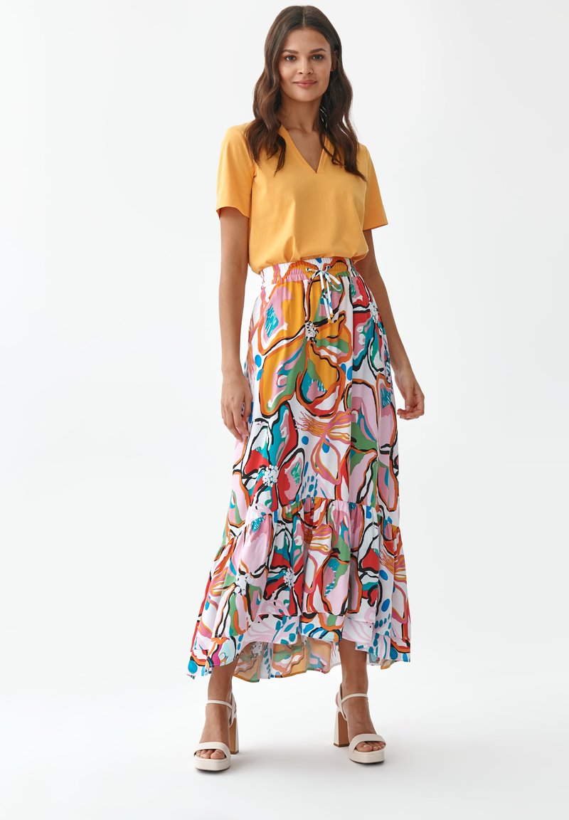 Żółta koszulka z dekoltem w kształcie litery V połączona z kolorową, wzorzystą maxi spódnicą z falbanami w odcieniach różu, zieleni, niebieskiego i pomarańczowego. Białe buty na blokowym obcasie.