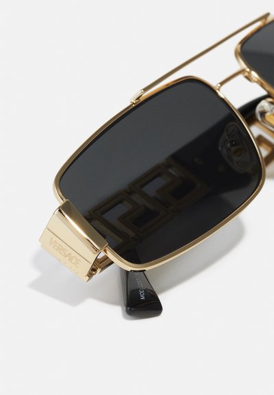 Ochelari de soare cu un cadru din metal auriu, lentile negre și un logo Versace pe lateral. Designul rectangular are brațe elegante și accente proeminente.