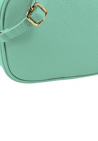 Kleine mintgroene leren tas met een gestructureerd oppervlak, gouden hardware en een vierkante gesp op de riem. Heeft afgeronde hoeken en nauwkeurige stiksels.