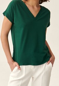 Vihreä V-kaula-aukkoinen t-paita, joka on valmistettu pehmeästä puuvillakankaasta, varustettuna käärityillä lyhyillä hihoilla ja rentoa istuvuutta, yhdistettynä valkoisiin housuihin.