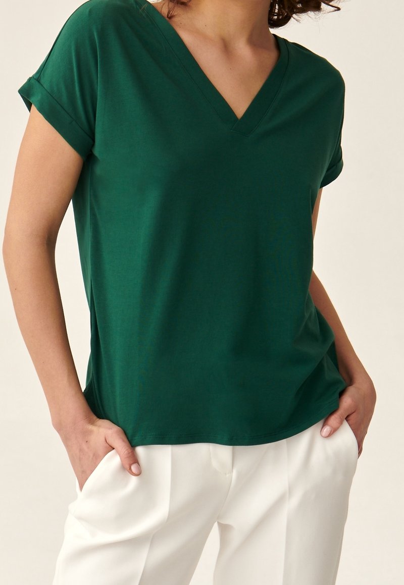 Vihreä V-kaula-aukkoinen t-paita, joka on valmistettu pehmeästä puuvillakankaasta, varustettuna käärityillä lyhyillä hihoilla ja rentoa istuvuutta, yhdistettynä valkoisiin housuihin.
