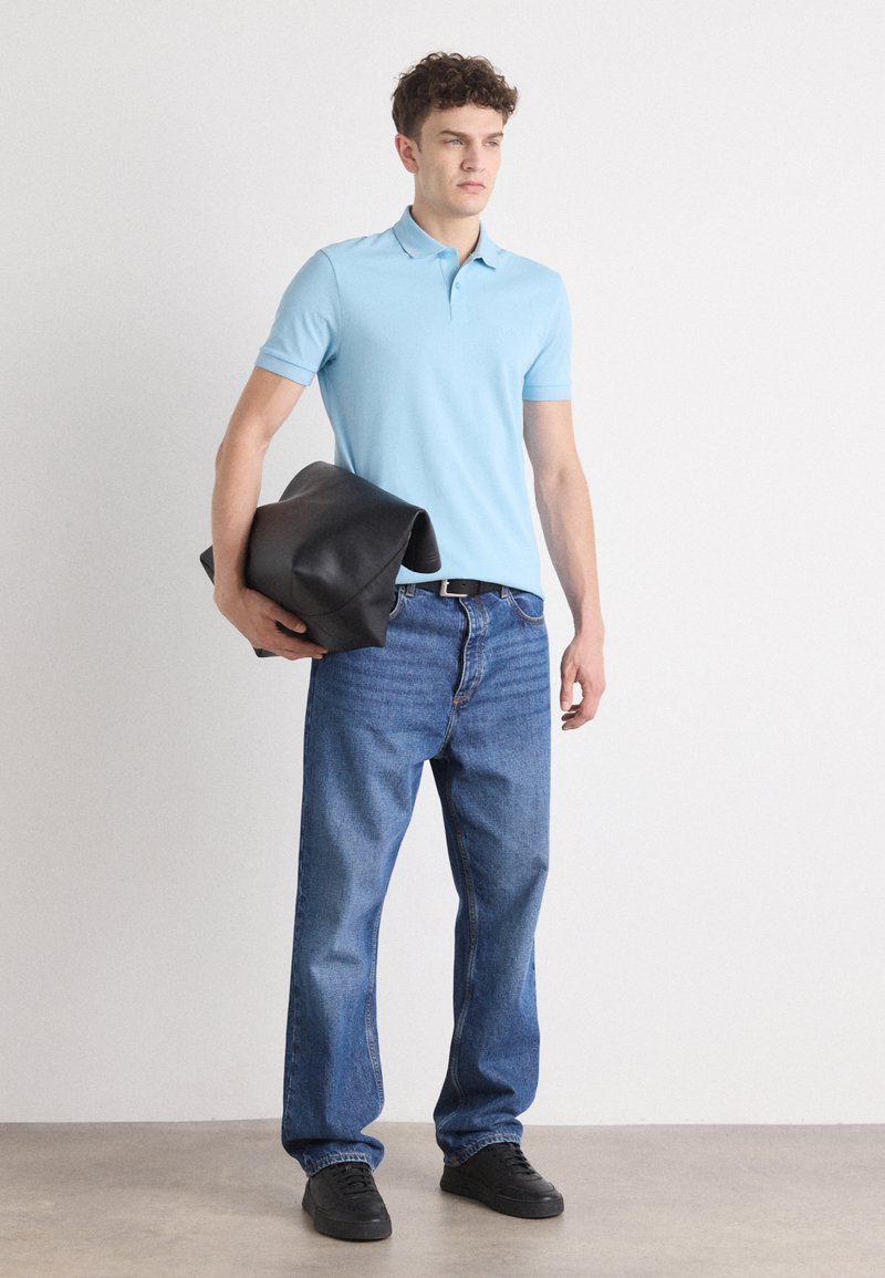 Lichtblauwe polo, donkerblauwe straight-leg jeans en zwarte sneakers. Houdt een zwarte leren tas met een gevouwen ontwerp vast.