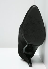 Zapato de tacón alto de ante negro con punta afilada y tacón de aguja estrecho; suela texturizada con detalles decorativos y marca.