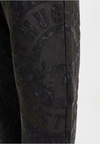 Mørkegrå joggebukser laget av et teksturert denim-lignende materiale. Merkbare svarte trykk inkluderer et hodeskallemotiv og "THUG LIFE"-logo med stjerner.