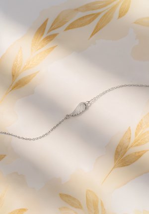 Bracelet en argent avec un élément central en forme d'aile, possédant une chaîne délicate et une texture lisse et poli sur un fond doux et texturé.