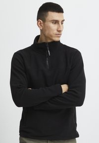 Sort fleece pullover med høj krave, frontlynlås og subtile stikninger. Lange ærmer med afslappet pasform og sidelommer.