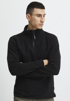 Black fleece pullover with a high collar, front zipper, and subtle stitching details. Long sleeves with relaxed fit and side pockets.