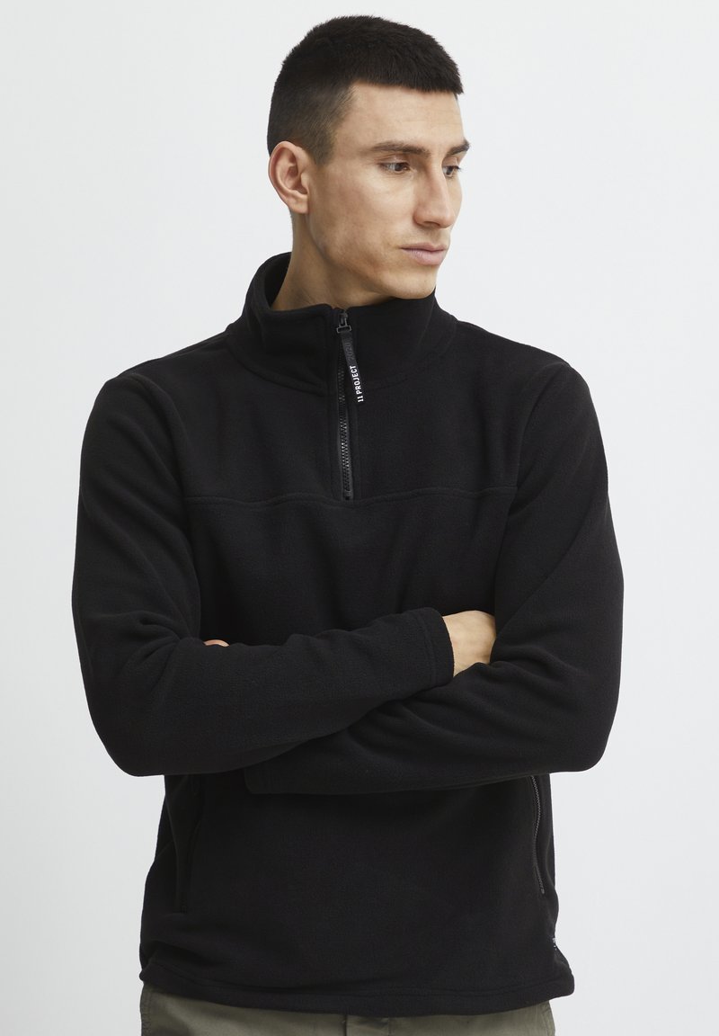 Svart fleece-pullover med høy krage, frontglidelås og subtile sømdetaljer. Lange ermer med avslappet passform og sidelommer.